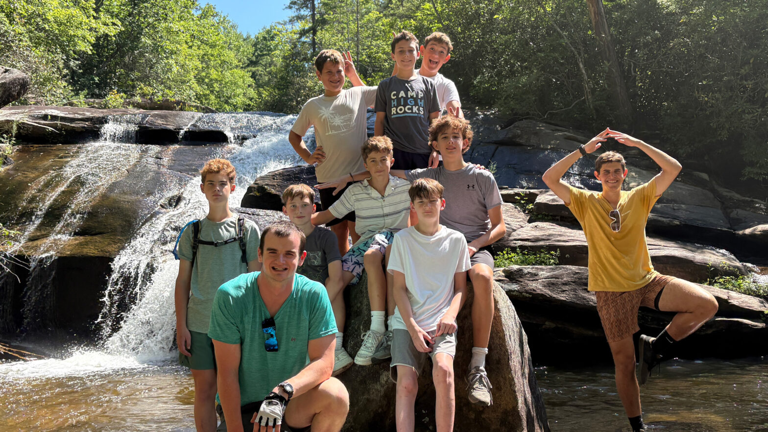 Group of boys posing on rocks beside a cascading waterfall in a forest, smiling and making playful gestures.