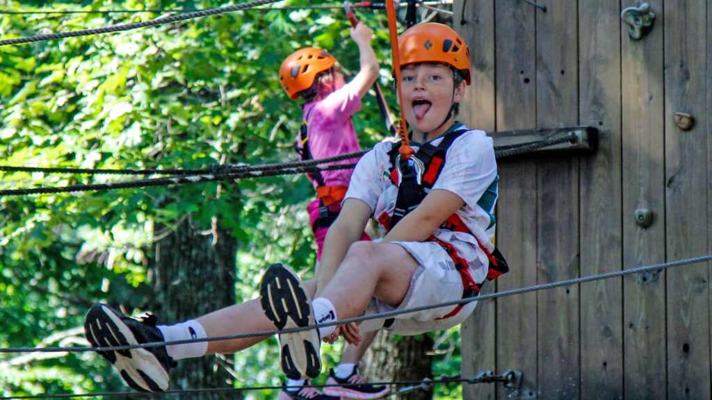 Two kids in orange helmets harnessed to safety lines, crossing a rope obstacle between trees and a wooden wall at a high-ropes course.