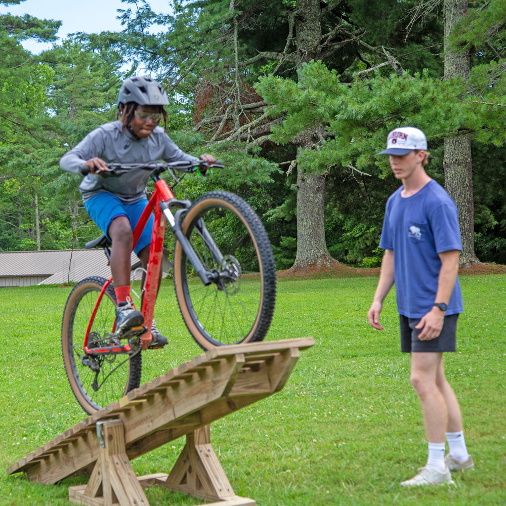 Rider wearing a helmet on a red mountain bike climbs a wooden ramp while a person watches on a grassy field.