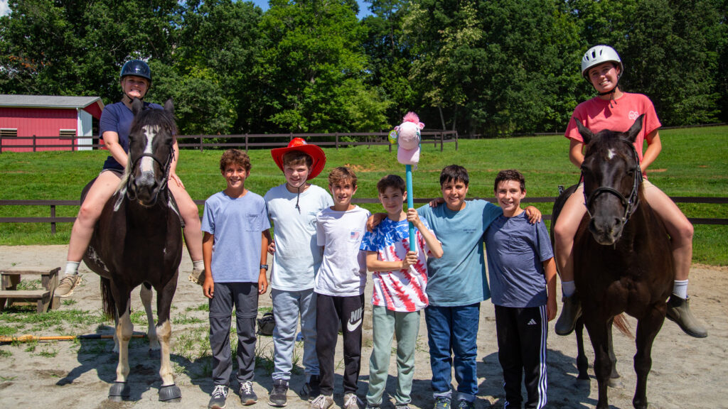 Group of kids standing together outdoors with two riders on horses at a riding range.