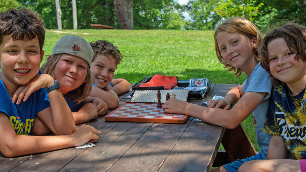 Group of children at a picnic table outdoors, playing chess on a checkered board in a sunny park.