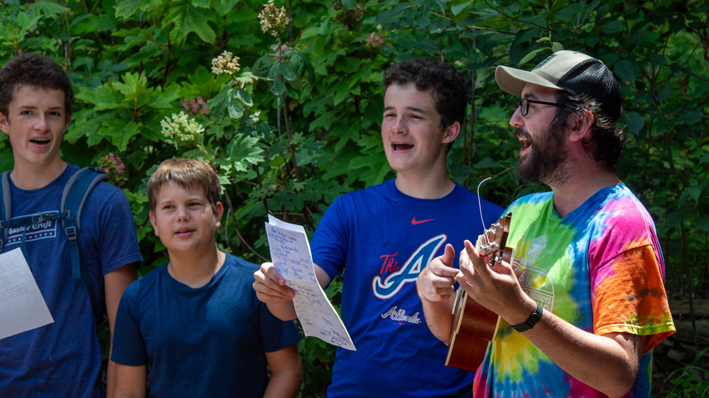 Group of four young men singing outdoors; one plays guitar while another holds sheet music.