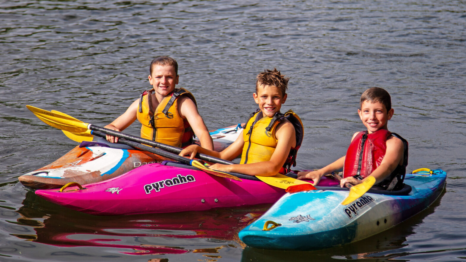 Three kids wearing yellow life jackets sit on colorful kayaks with paddles, smiling at the camera on a calm lake.