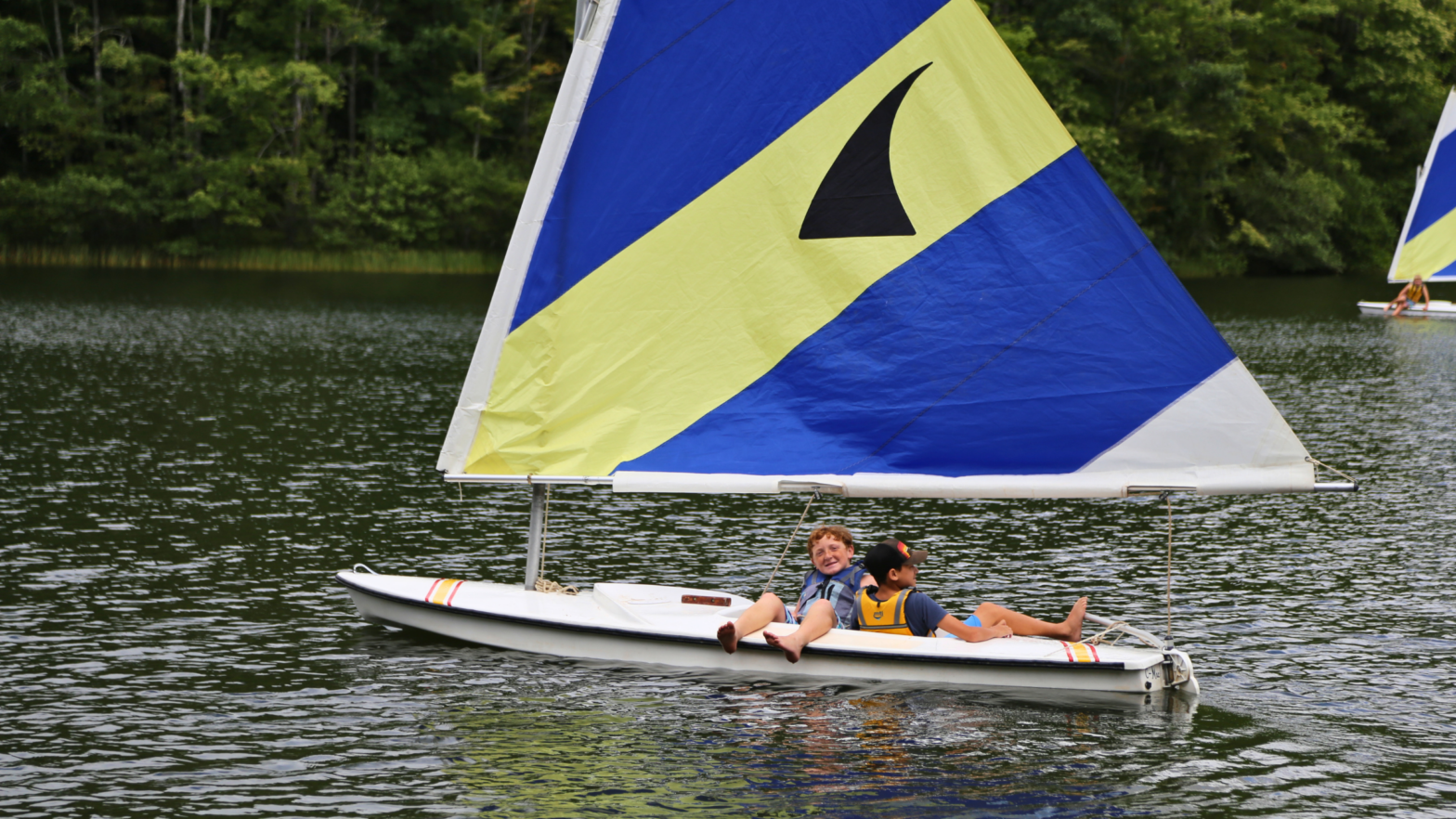 Two kids in life jackets lounging on a small sailboat on a calm lake, with a large blue-and-yellow sail dominating the scene and trees in the background.