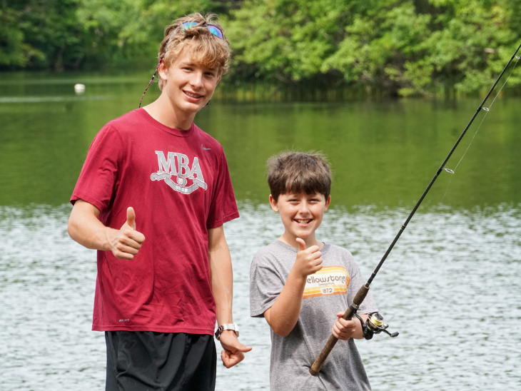 Older boy in a red 'MBA' shirt and younger boy in a gray tee hold a fishing rod by a lake, both giving thumbs up with smiles.