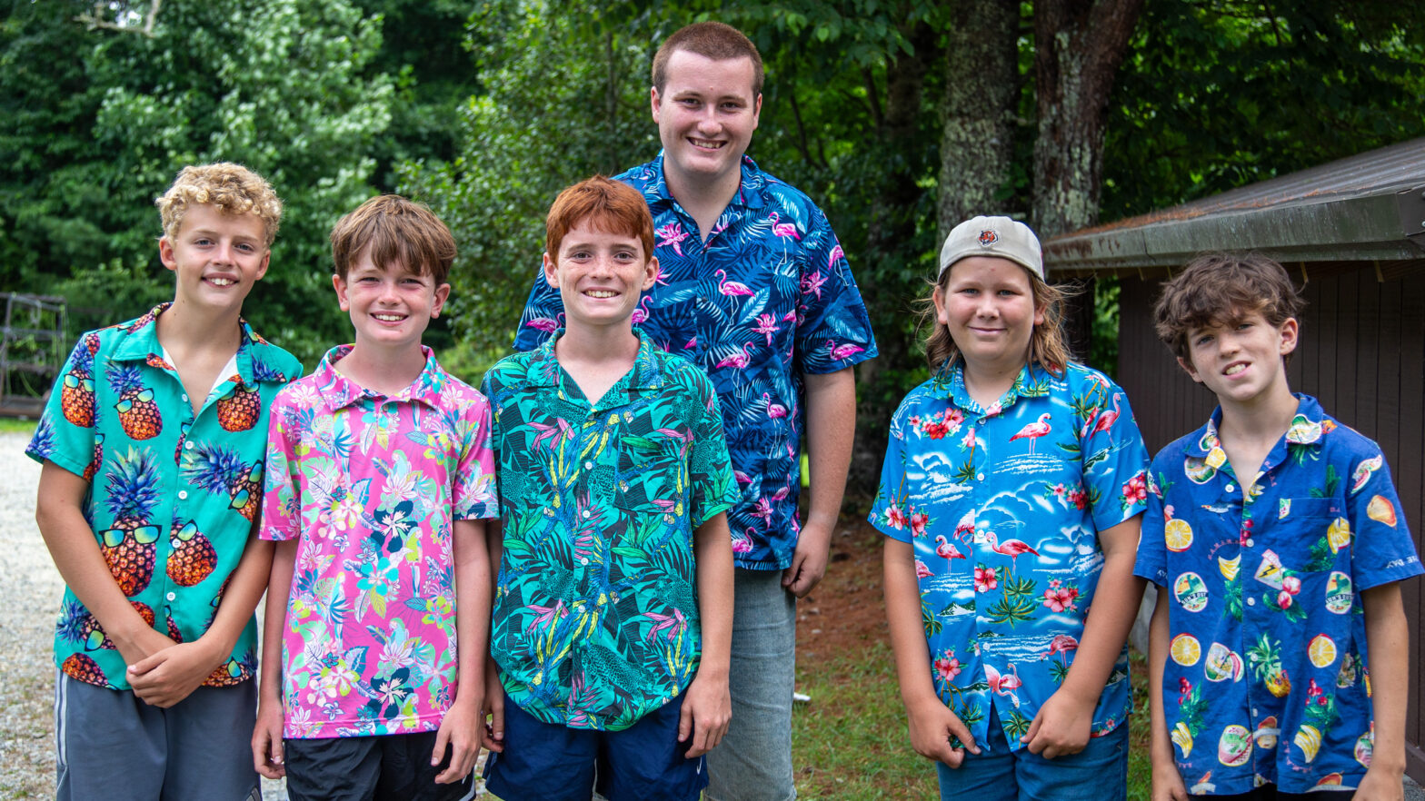 Five boys outdoors posing for a group photo in colorful Hawaiian shirts, with greenery in the background.