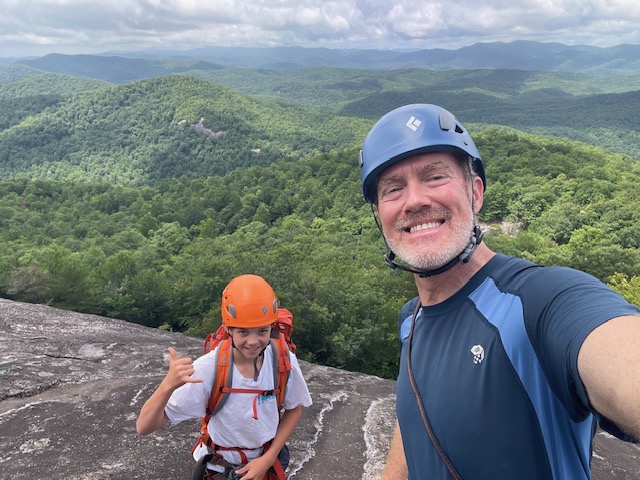 Two hikers wearing helmets atop a rocky ledge, posing for a selfie with forested mountains in the distance.
