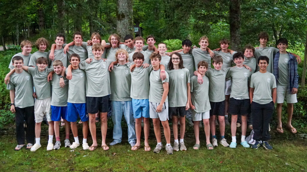 Large group of boys in matching gray shirts pose with arms around each other outdoors among trees.