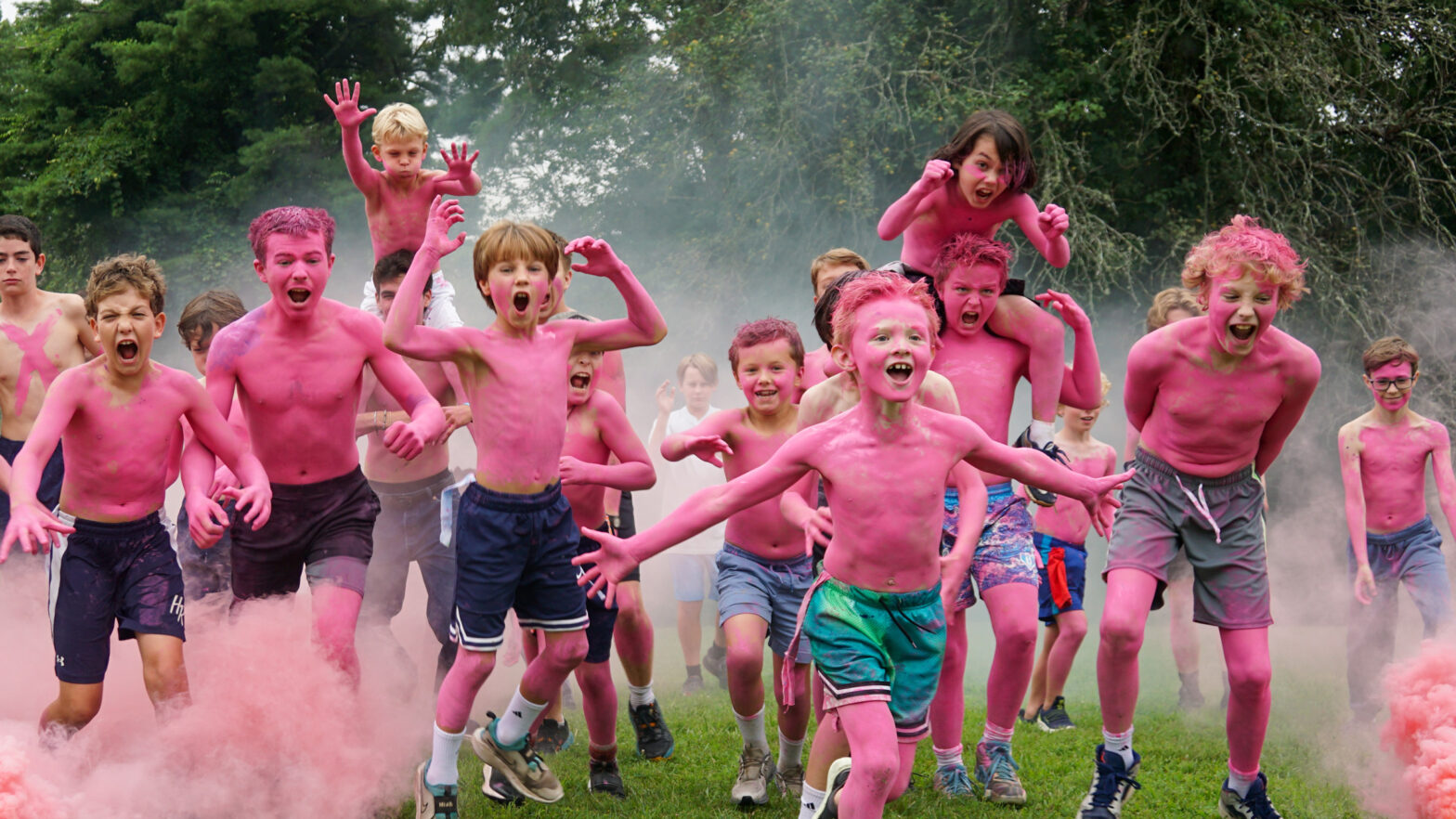 Group of shirtless kids sprinting through pink powder during a color run, outdoors on grass with trees in the background.