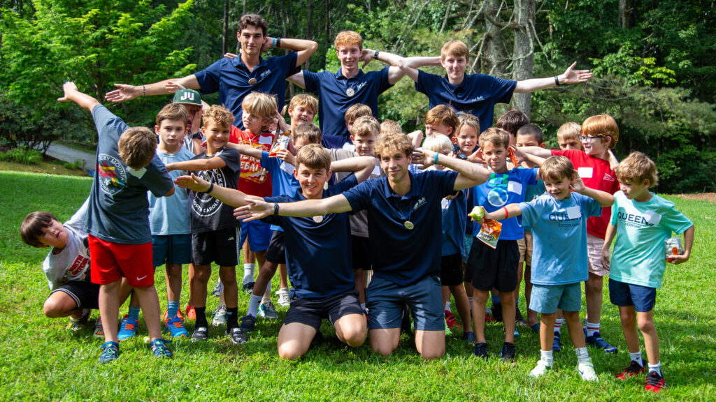 Large group of kids in a park posing with arms outstretched, many wearing matching navy camp shirts, smiling for a group photo.