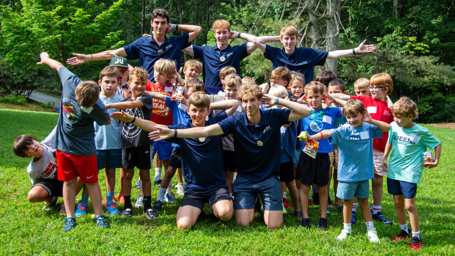 Large group of kids in a park posing with arms outstretched, many wearing matching navy camp shirts, smiling for a group photo.