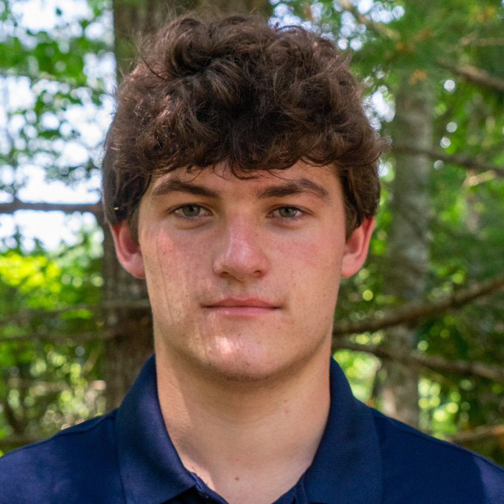 Young man with dark curly hair wearing a navy polo, standing outdoors with trees in the background.