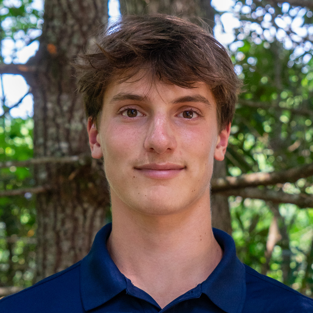 Head-and-shoulders portrait of a young man with brown hair wearing a navy polo, standing in a sun-dappled forest background.