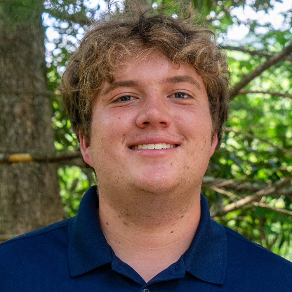 Smiling man with light brown hair outdoors among green trees, wearing a dark blue polo shirt.