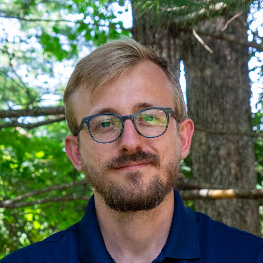 Man with blond hair and round glasses smiles outdoors, standing in a green forest backdrop with a tree behind him.
