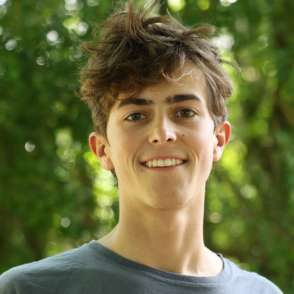 Young man with tousled brown hair smiles at the camera, outdoors among green foliage in a park setting.