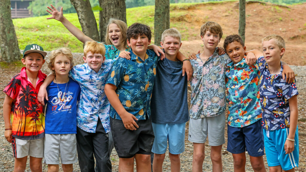 Group of kids in bright tropical shirts standing together outdoors, arms around each other and smiling in a park setting.
