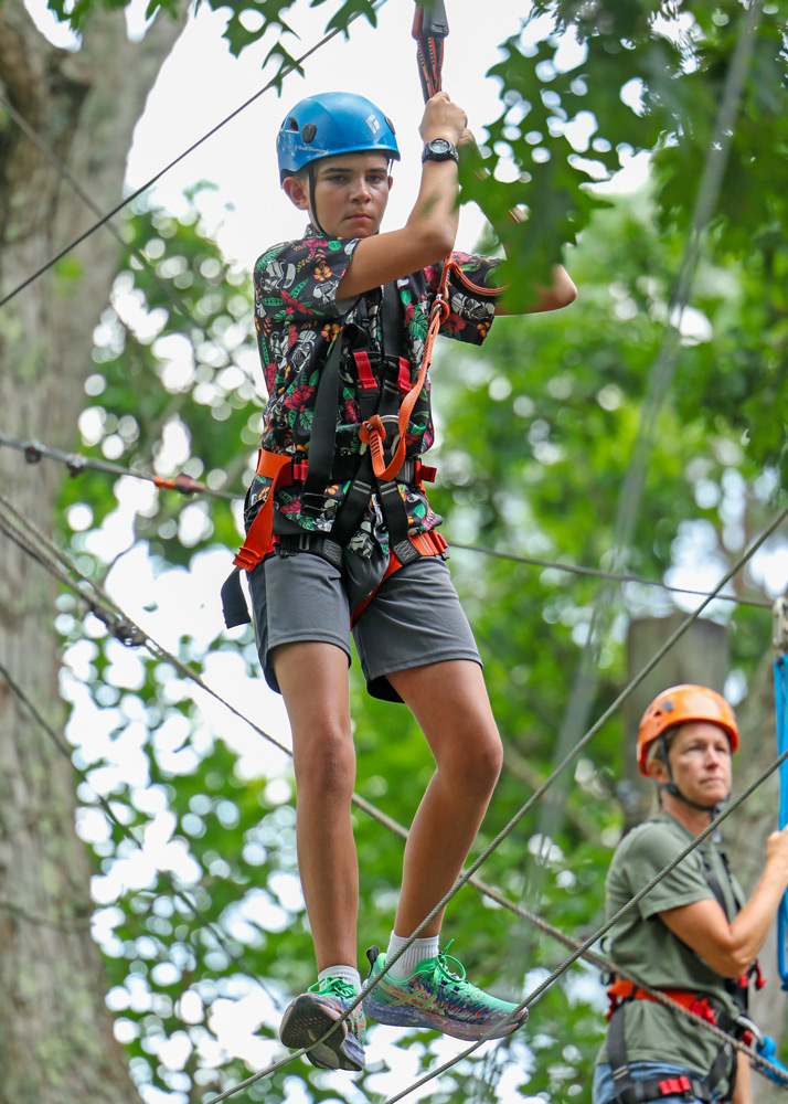 Child wearing a blue helmet and safety harness navigating a high ropes course among trees near greenery, outdoors.