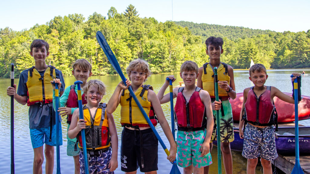 Group of children in bright life jackets holding paddles by a calm lake with green trees in the background behind them.