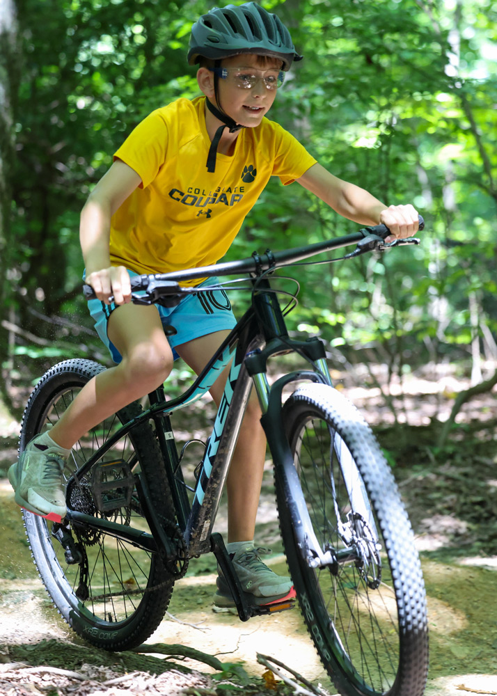 Child wearing a helmet and safety glasses rides a mountain bike through a sunny forest trail on a dirt path.