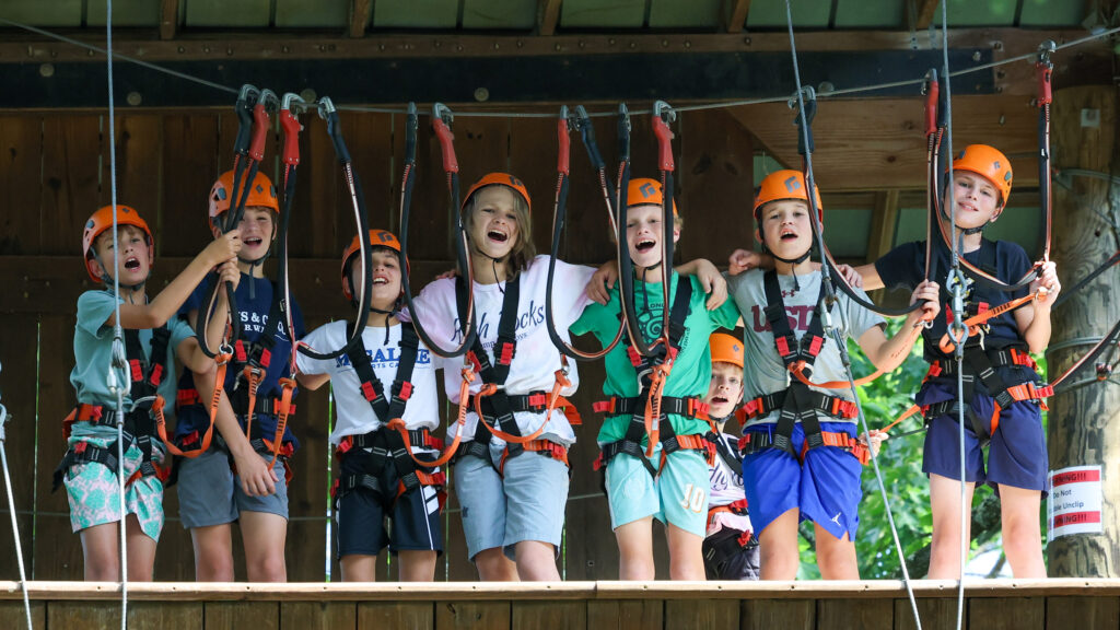 Group of children in orange helmets and harnesses on a wooden zip-lining platform ready for activity.