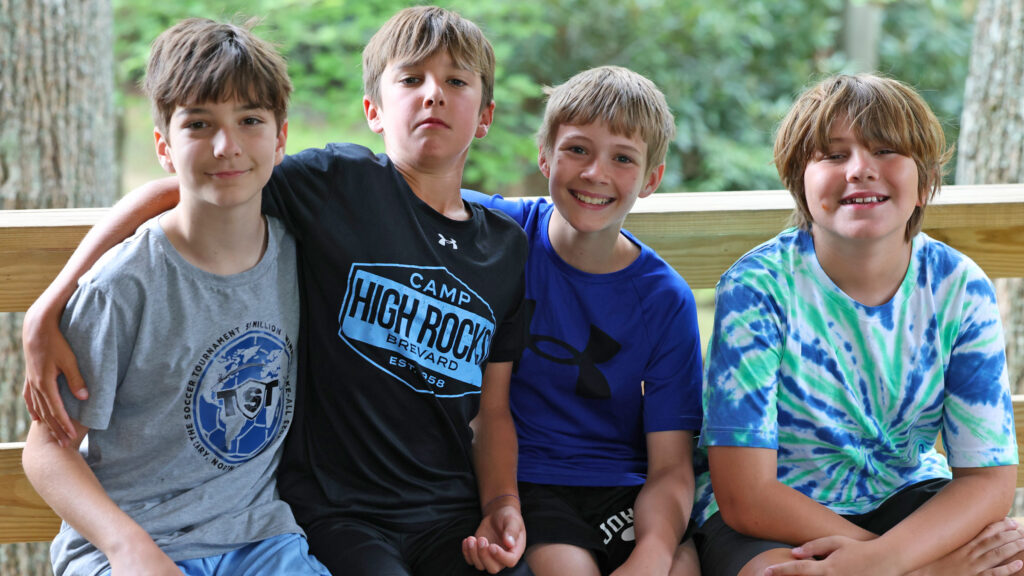 Four boys sit closely on a wooden bench outdoors, arms around each other, smiling at the camera.