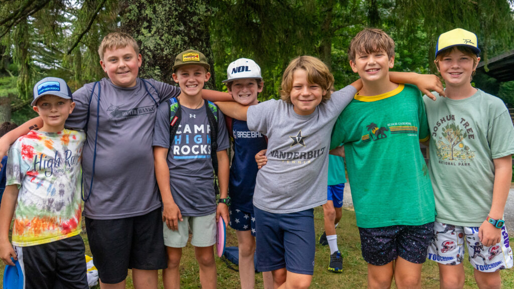 Group of smiling kids with arms around each other, posing outdoors at a camp or park, wearing colorful shirts and caps.