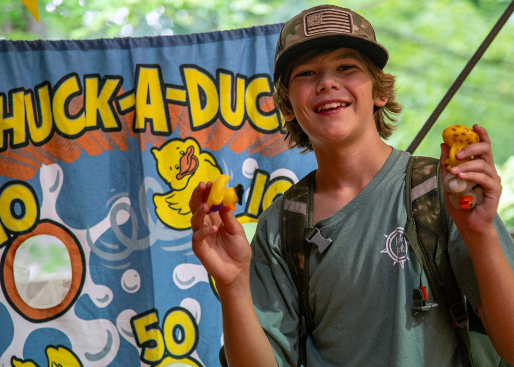 Smiling boy in a cap and backpack holding yellow rubber ducks at an outdoor Huck-A-Duck booth.
