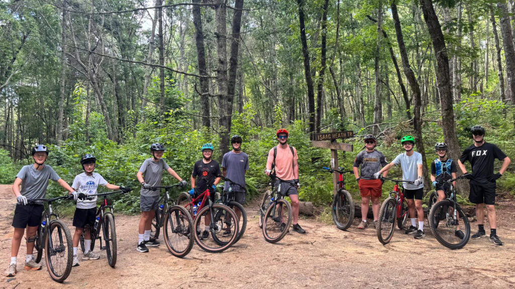 Group of ten cyclists with mountain bikes and helmets posing on a dirt forest trail, near a wooden signpost.