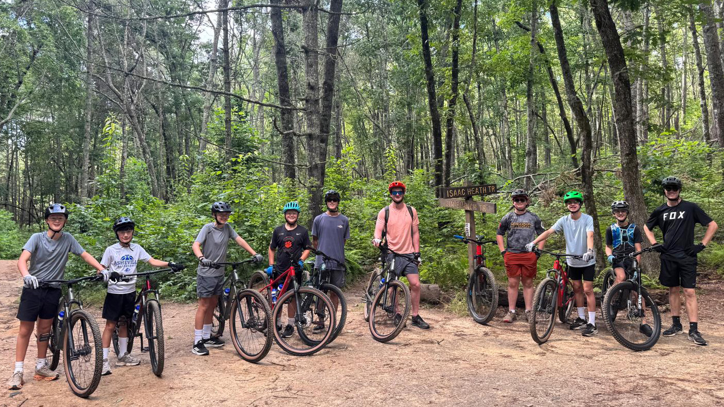 Group of ten cyclists with mountain bikes and helmets posing on a dirt forest trail, near a wooden signpost.
