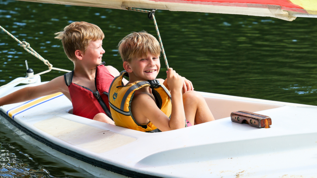 Two boys wearing life jackets sit in a small white sailboat on a calm green lake, smiling at each other.