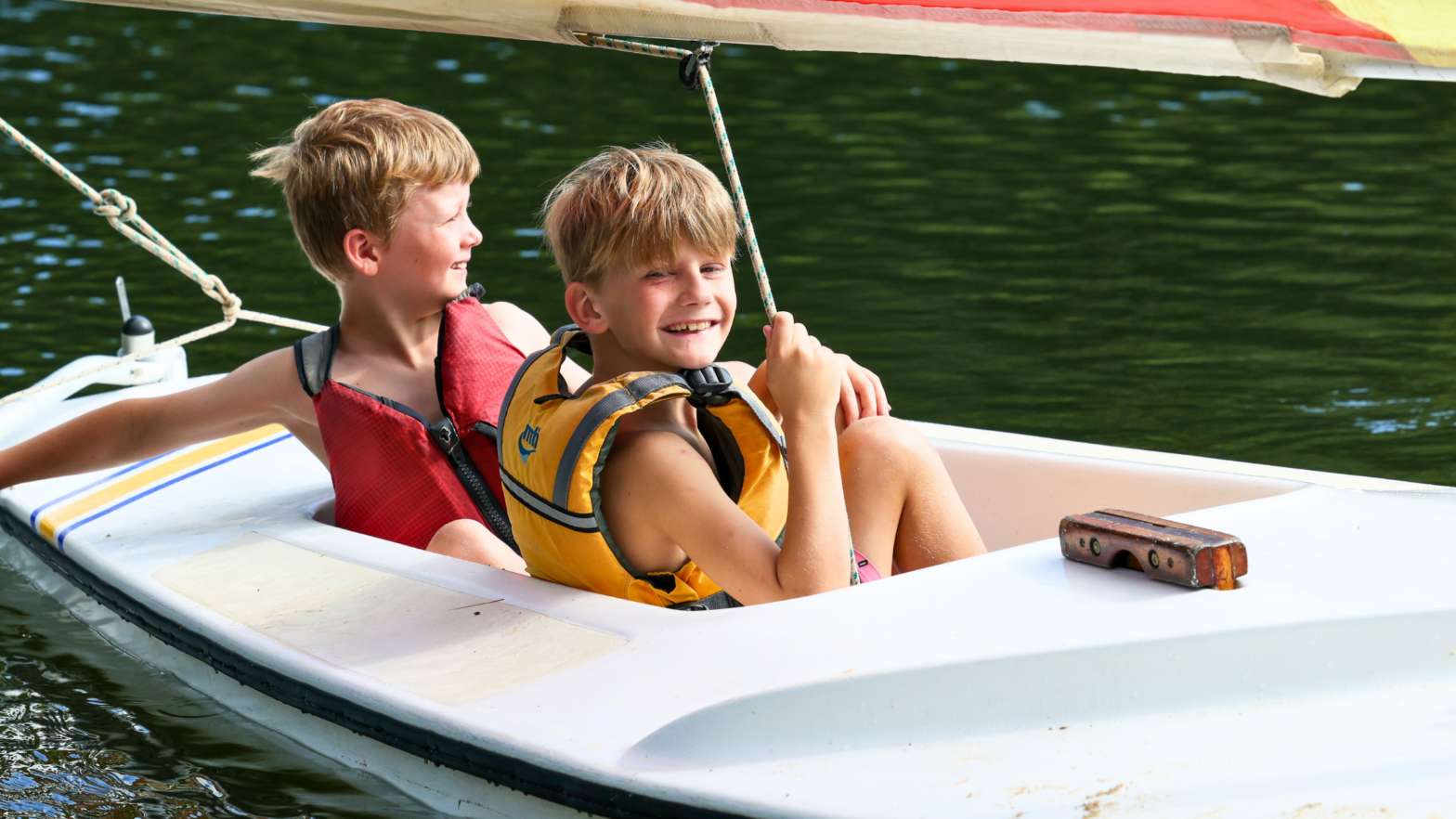 Two boys wearing life jackets sit in a small white sailboat on a calm green lake, smiling at each other.