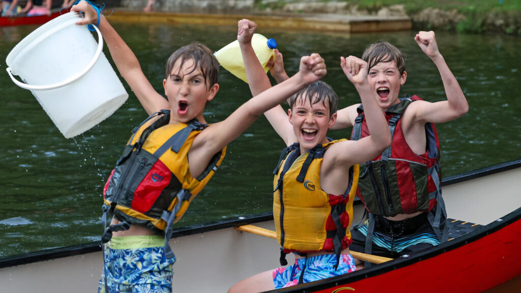 Three kids in life jackets cheer and raise their arms in a red canoe on the water.