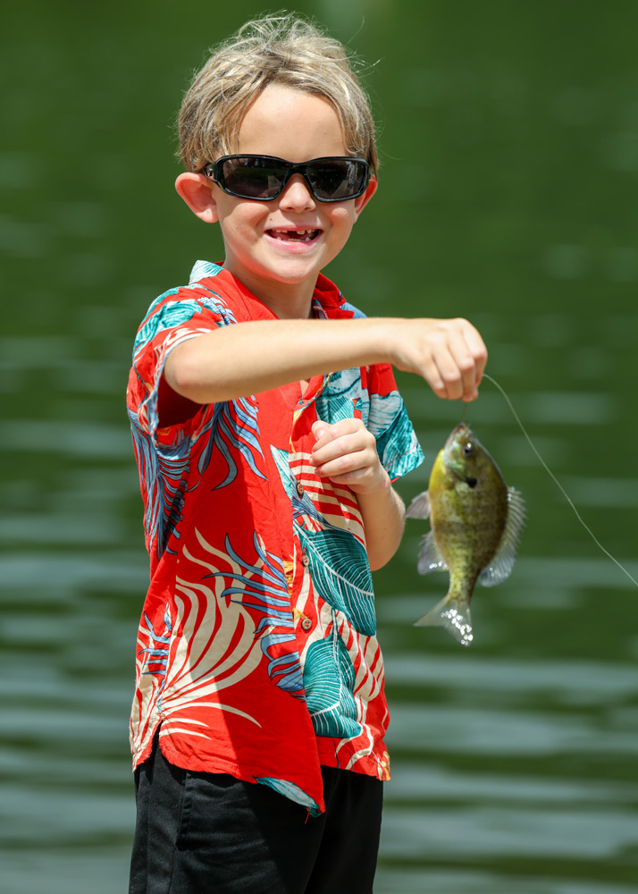 Young boy wearing sunglasses and a bright red tropical shirt shows off a small fish caught on a line by a green lake, smiling at the camera.