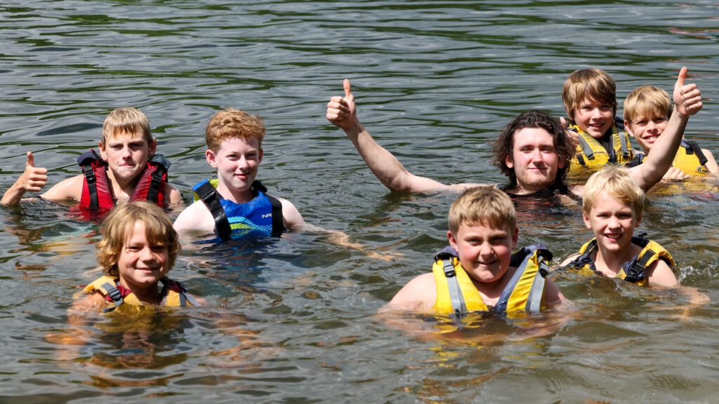 Group of children in life jackets swimming in a lake, giving thumbs up to the camera on a sunny day.