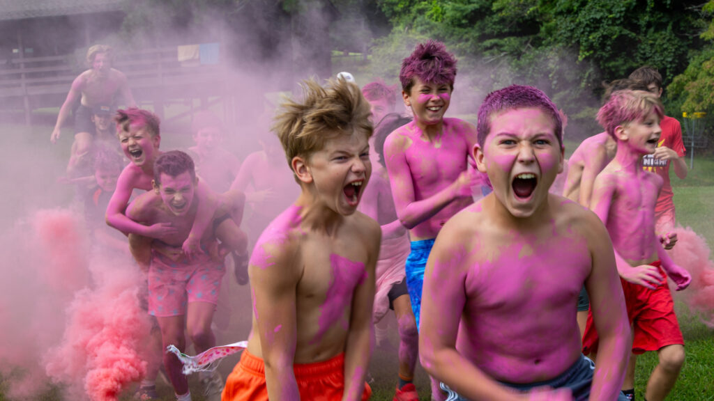 Group of shirtless kids running through pink powder at a color run, shouting and smiling outdoors.