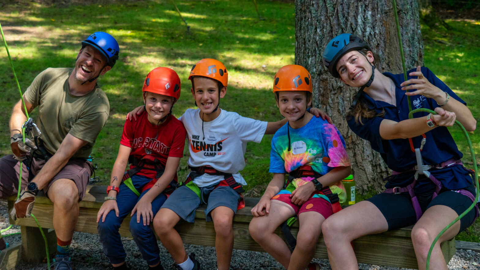 Five people wearing climbing helmets and harnesses sit on a wooden bench outdoors, smiling for the camera in a forest setting.