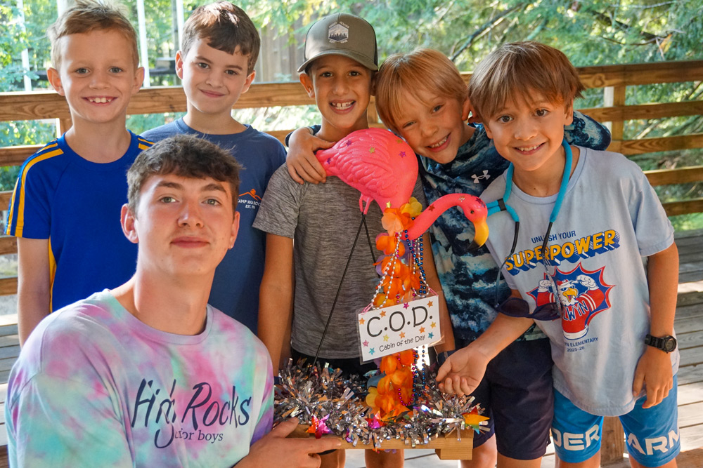 Six boys pose on a wooden deck, smiling, with a pink flamingo craft labeled 'COD: Cabin of the Day' in the center intent on a camp activity.