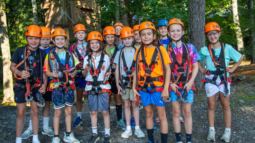 Group of children wearing orange helmets and climbing harnesses posing outdoors at a forest ropes course site