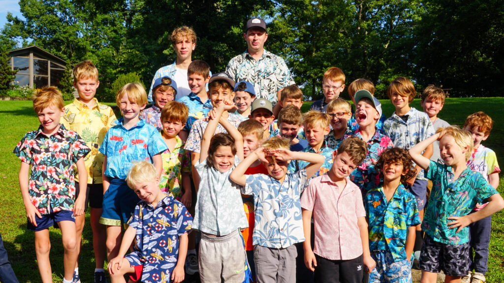 Group of children and a few adults posing outdoors in a sunny park, all wearing bright tropical Hawaiian shirts and shorts.