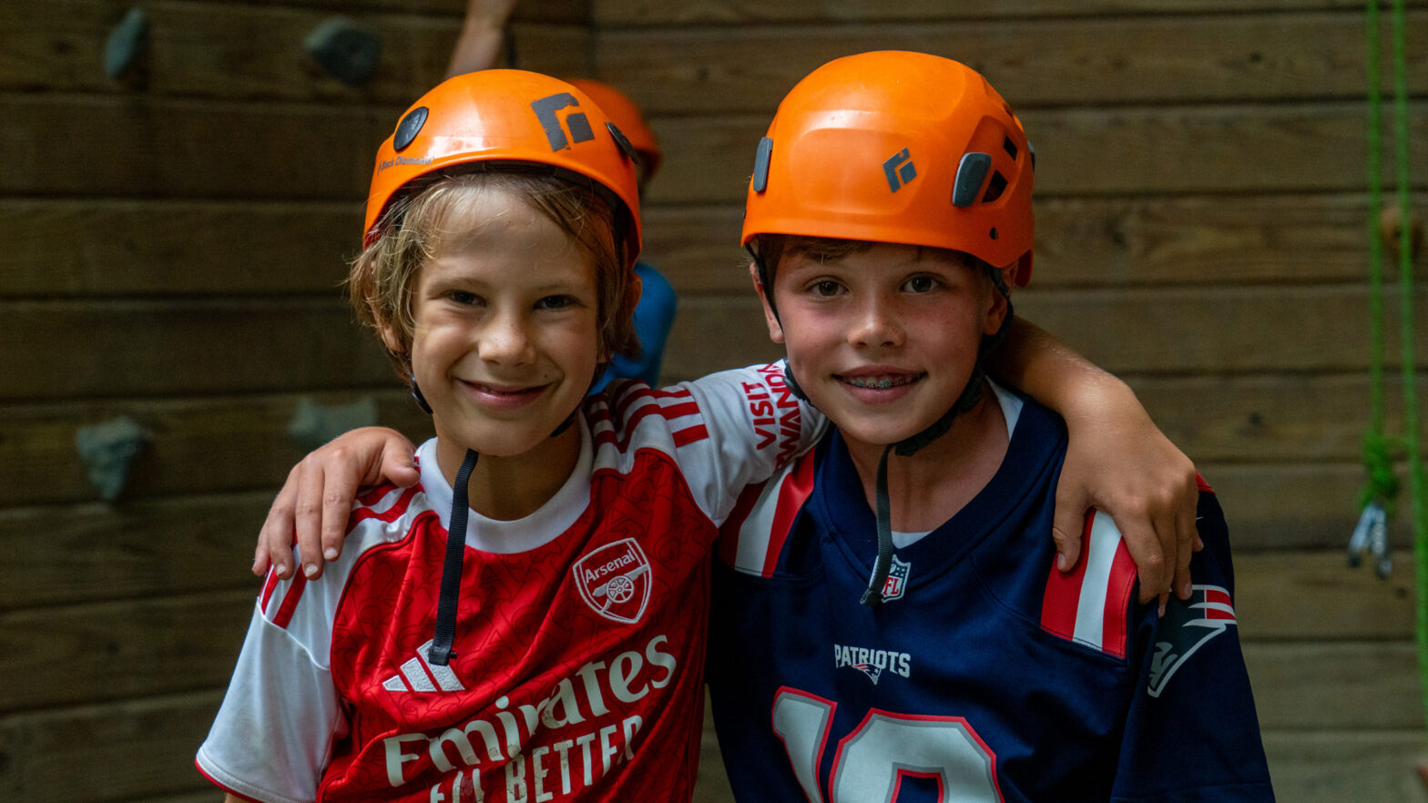 Two smiling kids with orange climbing helmets have their arms around each other in a climbing gym background.