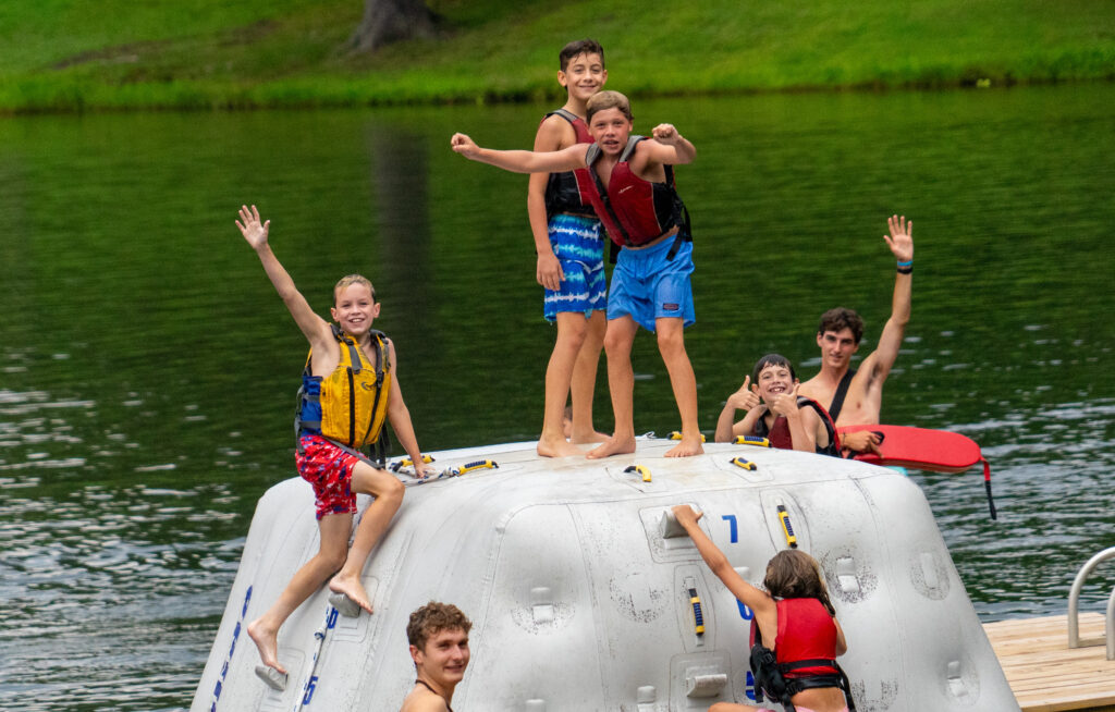 Children in life jackets on a large inflatable raft, waves and a green shoreline in the background, waving at the camera on a lake.