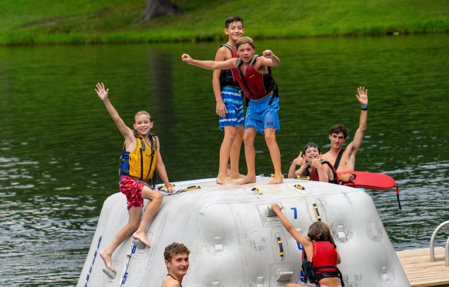 Children in life jackets on a large inflatable raft, waves and a green shoreline in the background, waving at the camera on a lake.